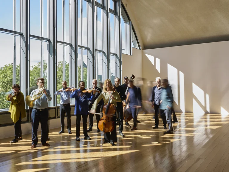Eine Gruppe von Musikern (Ensemble Modern) steht in einem gut beleuchteten Raum mit großen Fenstern. Sie haben verschiedene Musikinstrumente dabei.