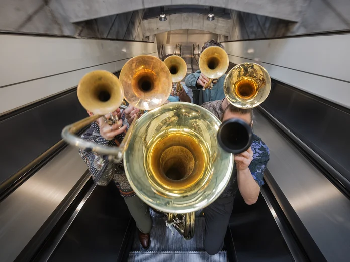 Eine Bläsergruppe (Federspiel) spielt auf einer Rolltreppe. Die Szene zeigt verschiedene Blechblasinstrumente in einem architektonischen Umfeld.