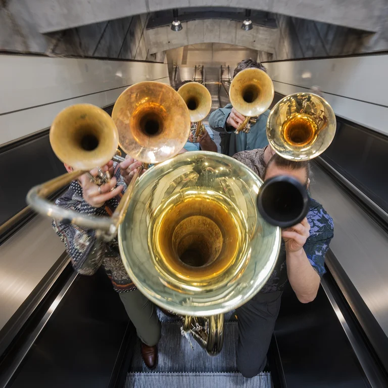 Eine Bläsergruppe (Federspiel) spielt auf einer Rolltreppe. Die Szene zeigt verschiedene Blechblasinstrumente in einem architektonischen Umfeld.