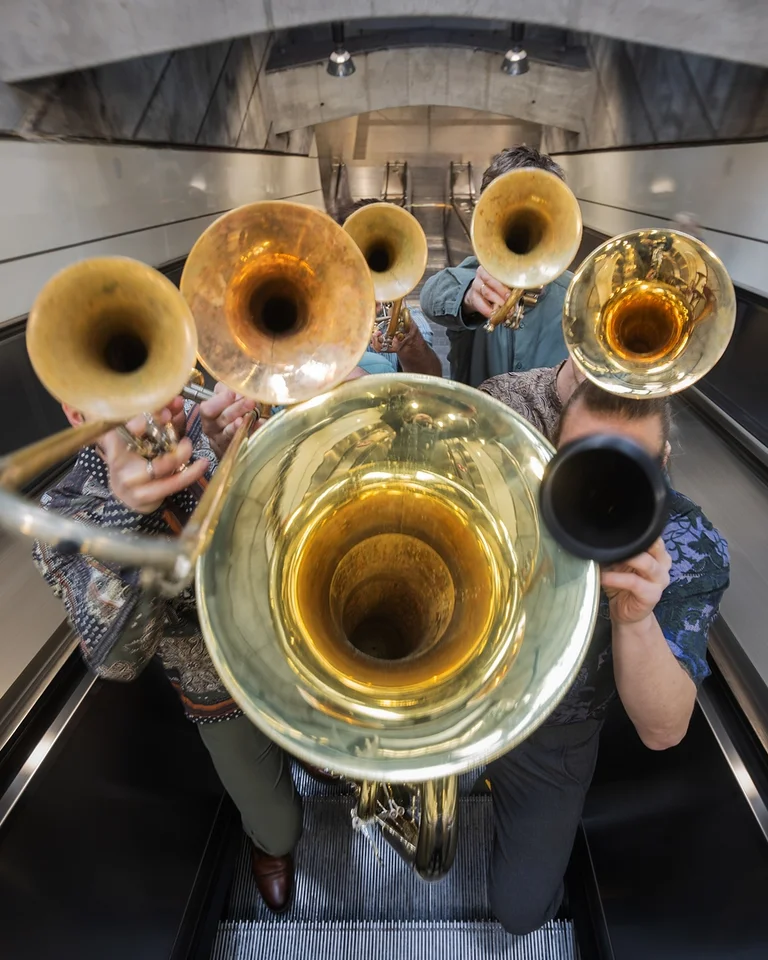 Eine Bläsergruppe (Federspiel) spielt auf einer Rolltreppe. Die Szene zeigt verschiedene Blechblasinstrumente in einem architektonischen Umfeld.
