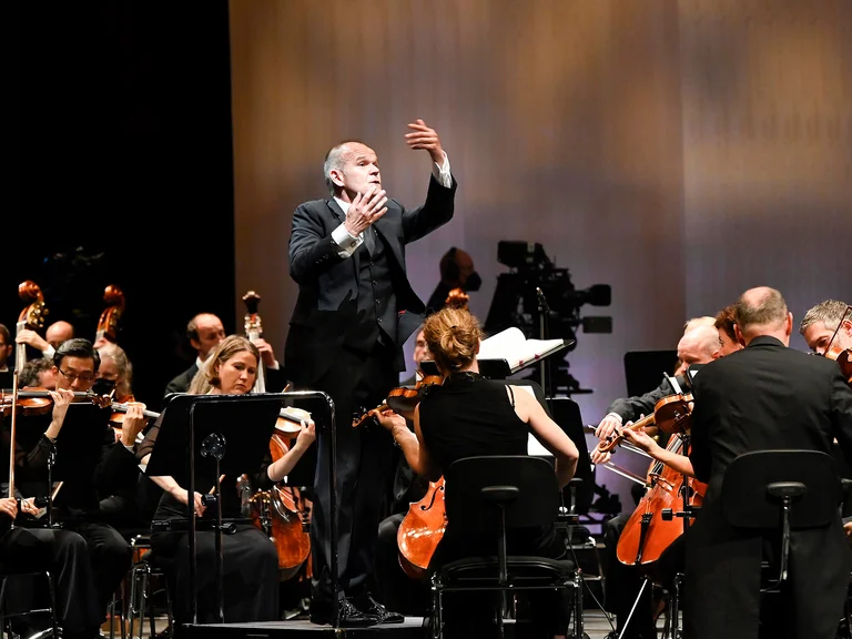 Ein Orchesterdirigent leitet eine Gruppe von Musikern während eines Live-Konzerts. Streicher sind gut sichtbar, das Ambiente ist beleuchtet.