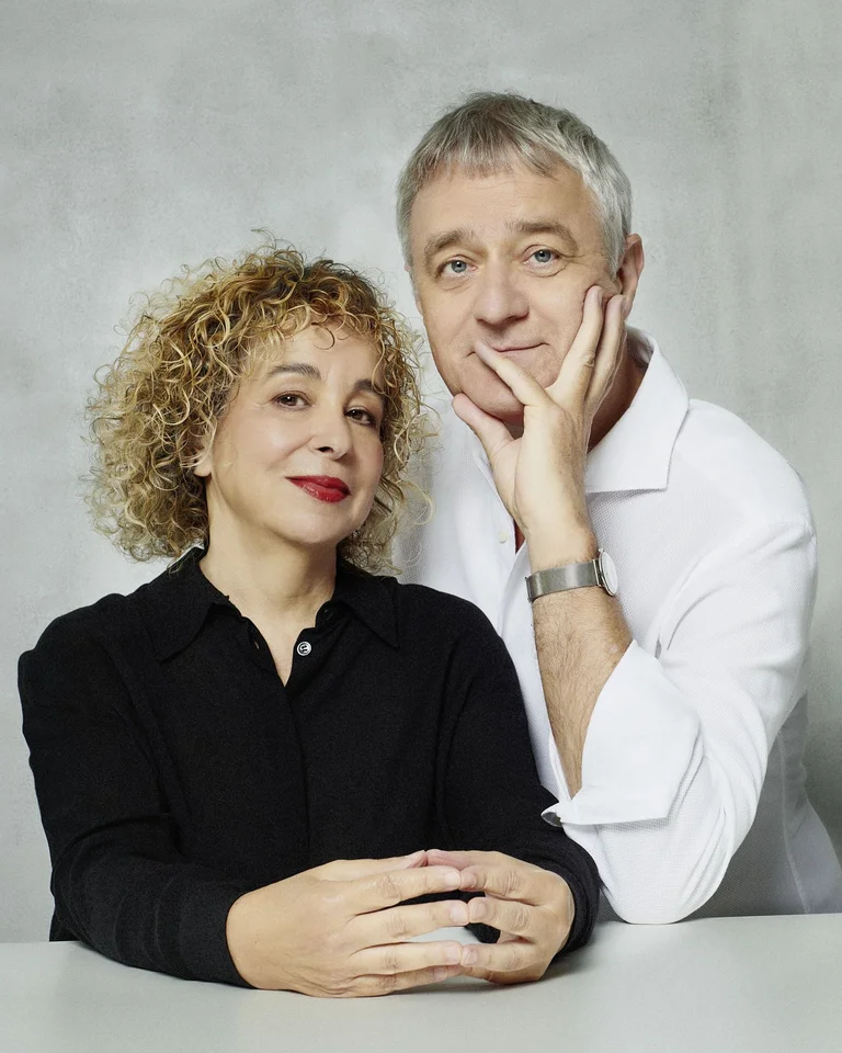 Zwei Personen in eleganter Kleidung posieren vor einem neutralen Hintergrund. Eine Frau mit lockigem Haar und ein Mann mit grauem Haar.