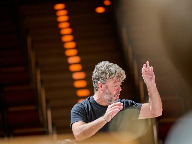 Ein Dirigent (Pablo Heras-Casado) mit lockigem Haar dirigiert konzentriert ein Orchester. Er erhebt eine Hand, während orange Lichter im Hintergrund leuchten.