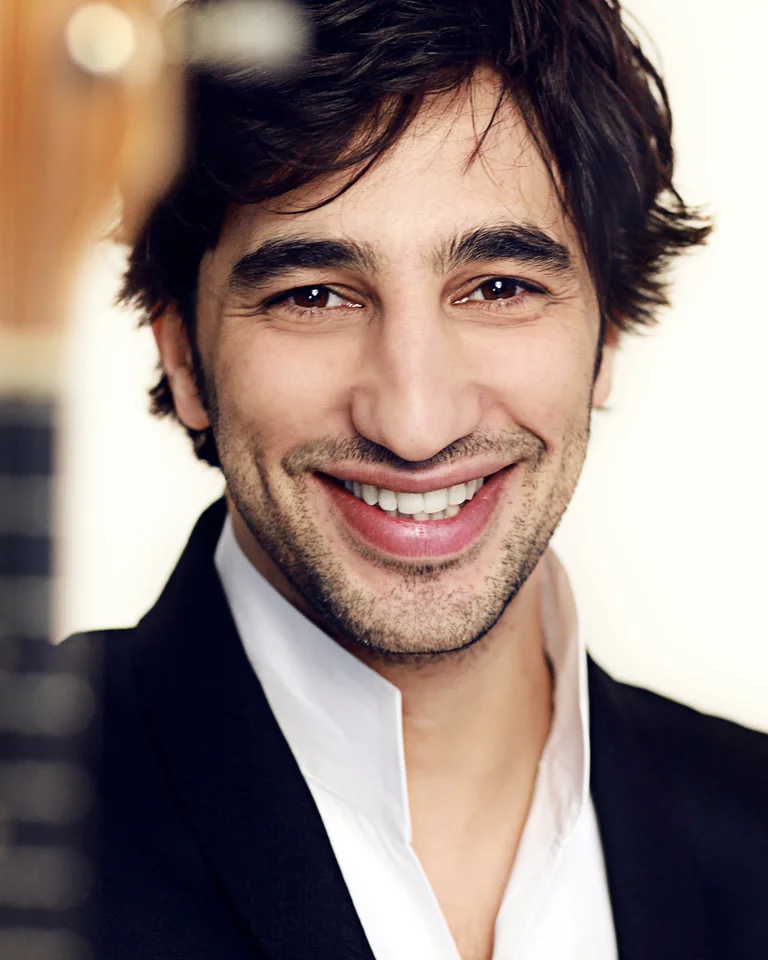 Lächelnder Mann in Anzug, im Vordergrund verschwommen ein Musikinstrument. Fröhliche Ausstrahlung, möglicherweise Musiker oder Künstler.