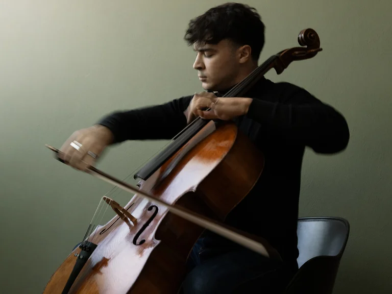 Ein Musiker (Petar Pejčić) spielt konzentriert Cello. Er sitzt auf einem Stuhl und der Hintergrund ist in weichen Grün- und Brauntönen gehalten.