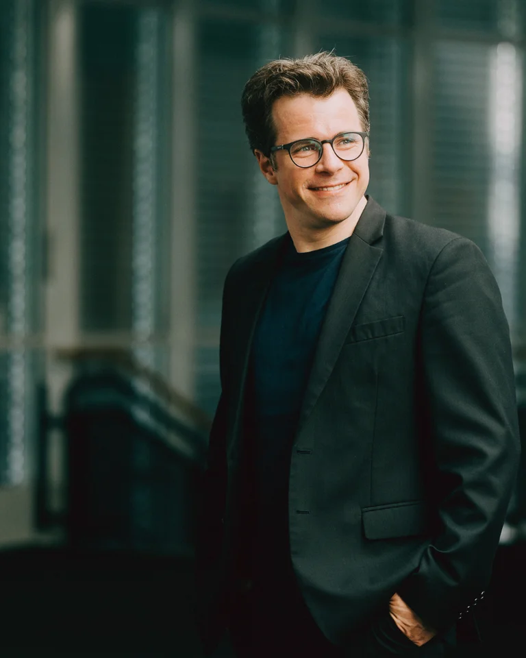 A man in a black suit is smiling while standing in a modern building with glass walls and an urban backdrop.