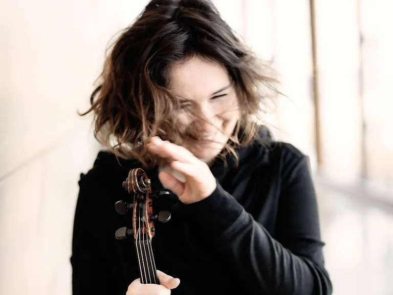 Eine Frau (Patricia Kopatchinskaja) mit welligem Haar hält eine Geige und lächelt. Sie trägt dunkle Kleidung und der Hintergrund ist unscharf.